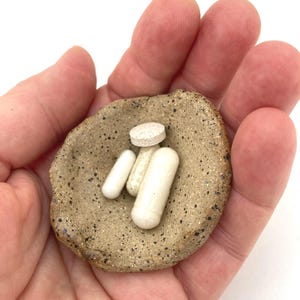 May include: A small, round, speckled tan ceramic dish holding three white pills. The dish has a textured, earthy appearance with dark brown flecks scattered across its surface. The pills consist of two elongated capsules and one flat, round tablet.