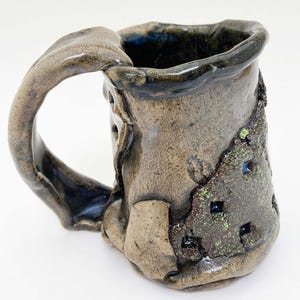 May include: Handmade ceramic mug with a unique, textured design. The mug features a large, organically shaped handle and a rough, earthy exterior with dark green and blue accents. The mug's interior is a deep, glossy blue.