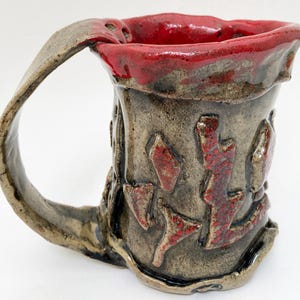 May include: Handmade ceramic mug with a textured, earthy brown exterior and a vibrant red interior. The mug features raised, abstract red designs and a large, curved handle. A unique piece for enjoying beverages.
