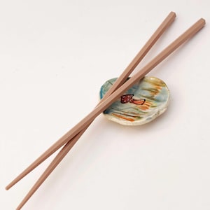 May include: A pair of light brown chopsticks resting on a small ceramic dish with a mushroom design. The dish has a blue, green, and orange glaze.