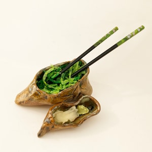 May include: A ceramic bowl shaped like a shoe, filled with green seaweed salad and two black chopsticks with green and white floral designs.