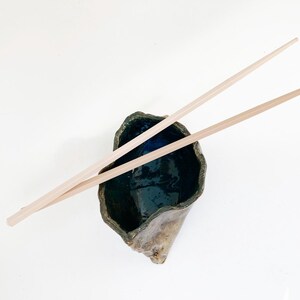 May include: A handmade ceramic bowl with a dark blue interior and a textured, organic exterior. Two light-colored wooden chopsticks rest across the bowl, suggesting a culinary theme. The bowl's unique shape and color contrast create visual interest.