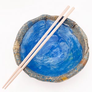 May include: A round, handcrafted ceramic bowl with a vibrant, glossy blue interior and a textured, mottled brown and blue exterior rim. A pair of smooth, light-colored wooden chopsticks rests diagonally across the top of the bowl.
