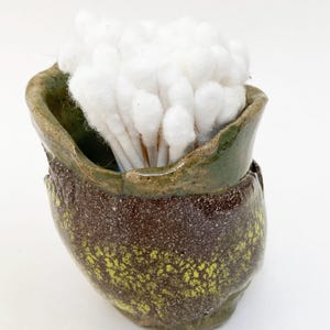 May include: A handmade ceramic container with a dark brown base and green rim, filled with white cotton swabs. The container has a textured, speckled design with yellow accents. The design is organic and rustic.