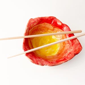 May include: A handmade ceramic bowl with a vibrant red, orange, and yellow gradient. Two light-colored wooden chopsticks rest across the bowl. The bowl's design suggests a culinary theme, perfect for serving Asian-inspired dishes.