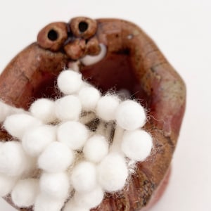 May include: A ceramic holder shaped like a face with a wide open mouth, holding a bunch of white cotton swabs.