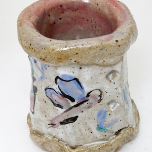 May include: A small, white ceramic vase with a pink rim and a hand-painted design of blue and pink flowers. The vase has a rough, textured surface and a slightly uneven rim.