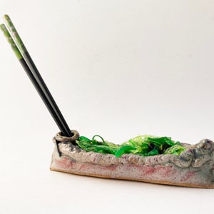 May include: A ceramic chopstick holder shaped like a boat with green and white accents. The holder has a ring for holding chopsticks and a green and white design.