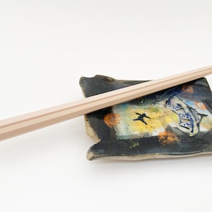 May include: A pair of light-colored wooden chopsticks resting on a small, square ceramic dish. The dish features a dark background with a colorful abstract design and a small figure. The chopsticks are approximately 9 inches long.