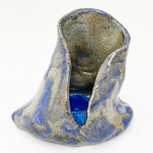 May include: A handmade ceramic sculpture with a unique, organic shape. The piece features a blend of blue, gray, and tan glazes, creating a textured appearance. The interior has a vibrant blue hue. The sculpture is a decorative art piece.