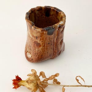 May include: A small, brown ceramic vase with a rough, textured surface. The vase has a wide opening and a slightly irregular shape.