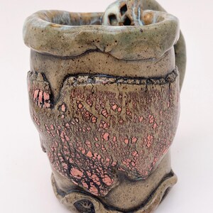 May include: A ceramic mug with a textured, brown and green glaze. The mug has a unique, sculpted handle and a copper-colored glaze on the side.
