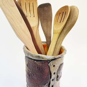 May include: A collection of wooden kitchen utensils, including spatulas and slotted spoons, displayed in a decorative ceramic utensil holder. The holder is a mix of brown, red, and beige tones, with a textured surface and unique design.