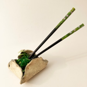 May include: A ceramic bowl with a rough, textured surface holds green seaweed and two black chopsticks with green and pink floral designs.