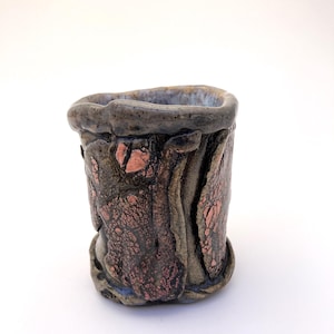 May include: A small, cylindrical ceramic cup with a textured, brown and copper glaze. The cup has a slightly irregular rim and a rough, uneven surface.