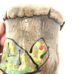 May include: A handmade ceramic mug with a textured, earthy brown exterior. The mug features a colorful abstract design with green, yellow, and red accents outlined in black. The rim has a unique, uneven texture.