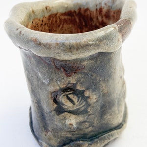 May include: A small, gray ceramic pot with a brown rim and a unique design. The pot has a rough, textured surface and a slightly irregular shape.