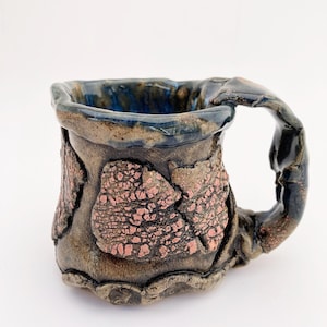 May include: A handmade ceramic mug with a blue glaze and copper accents. The mug has a unique, irregular shape and a textured surface.