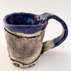 May include: Handmade ceramic mug with a textured, layered design. The mug features a dark blue rim and handle, with a textured, purple-brown band around the middle. The base is a lighter, silver-grey color with embossed star-like shapes.