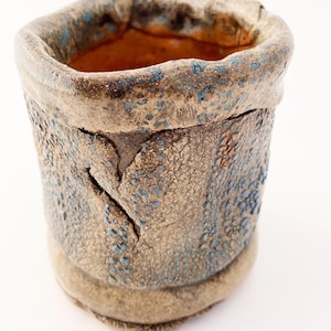May include: A handmade ceramic vessel with a textured, speckled surface. The exterior features a blend of tan and brown hues with blue accents. The interior has an orange-brown color. The cylindrical shape has a slightly irregular rim and a crack on the side.