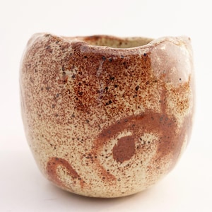 May include: A small, rounded ceramic cup with an irregular, wavy rim. The exterior features a speckled, cream-colored base glaze overlaid with textured, reddish-brown splatters and swirling, hand-painted abstract patterns across the surface.