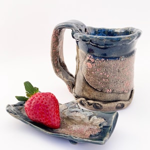 May include: A handmade ceramic mug with a matching small plate. The mug is primarily gray with blue and pink accents, and a large handle. A fresh red strawberry rests on the plate. The mug and plate are on a white background.