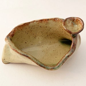 May include: A small, off-white ceramic bowl with a speckled glaze and a small, rounded spout. The bowl has an irregular shape and a slightly textured surface.