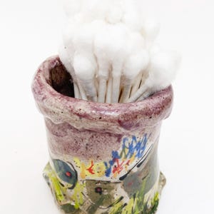 May include: A small, hand-painted ceramic container with a purple rim and colorful abstract designs. The container is filled with white cotton swabs, creating a functional and artistic bathroom accessory.