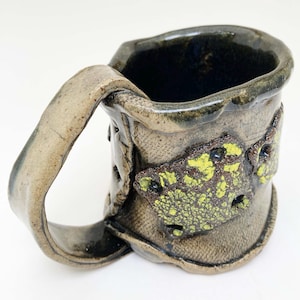 May include: A handmade ceramic mug with a textured, speckled brown glaze. The mug has a unique, asymmetrical shape and a large handle. The mug features a decorative design of two yellow and brown butterflies.