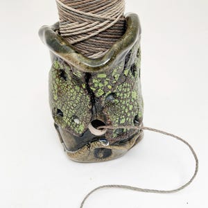 May include: A handmade ceramic twine dispenser. The dispenser is a mottled green and brown color with a textured surface and small holes. A spool of twine sits inside, with the end of the twine extending out of the dispenser.