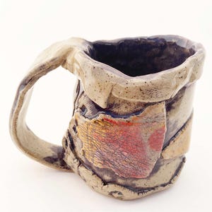 May include: Handmade ceramic mug with a unique, textured design. The mug features a tan handle and body with patches of red, yellow, and orange. The interior is dark, and the overall aesthetic is rustic and artistic.