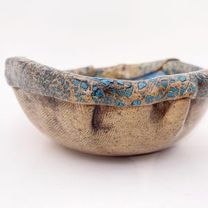 May include: A small, round ceramic bowl with a textured, tan-colored exterior featuring a cross-hatched pattern. The thick, rolled rim is finished with a crackled, dark brown and bright turquoise glaze that contrasts with the neutral body of the bowl.