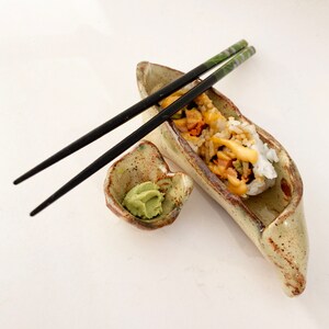 May include: A ceramic dish shaped like a pea pod with a single sushi roll inside. The dish is brown and green with a speckled glaze. A small dish with green wasabi paste sits next to the pea pod. A pair of black chopsticks are laying on top of the dish.