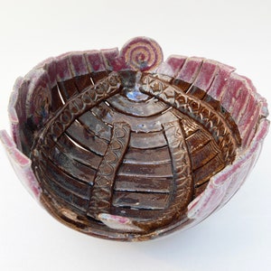 May include: A ceramic bowl with a glossy dark brown interior featuring embossed geometric patterns and a scalloped rim glazed in a muted mauve purple. The exterior edge displays a small, raised spiral detail at the top center of the rim.