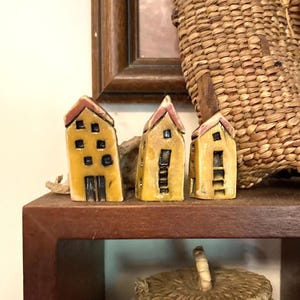 May include: Three small ceramic house figurines in a mustard yellow glaze with dark brown accents. Each house features a peaked roof, rectangular windows, and a door. The structures are arranged in a row, showcasing a textured, hand-crafted finish.