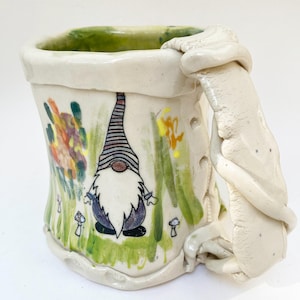 May include: A handmade ceramic mug with a whimsical gnome design. The mug features a white base with a green interior and a textured handle. The exterior is decorated with a gnome, mushrooms, and colorful floral accents. The mug is approximately 4 inches tall.