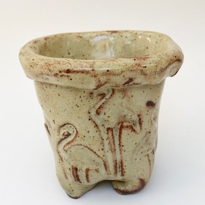May include: A small, light green ceramic planter with a brown glaze. The planter has a rounded base and a wide, flared rim. The sides of the planter are decorated with a raised design of stylized birds.