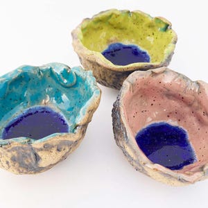 May include: Three small ceramic bowls with a textured, rough exterior. Each bowl has a different colored glaze on the outside: teal, yellow, and pink. The inside of each bowl is glazed with a dark blue color.