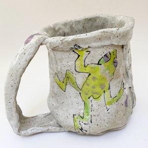 May include: A handmade ceramic mug with a speckled off-white glaze and an irregular, textured rim. The exterior features a hand-painted illustration of a bright green frog with purple spots and black outlines, complete with a thick, sturdy handle.