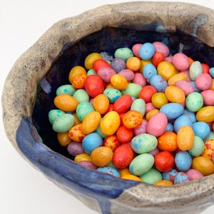 May include: A ceramic bowl with a blue glaze and speckled brown exterior, filled with colorful candy-coated chocolate eggs.