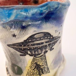 May include: A ceramic mug with a unique design featuring a UFO and a person being beamed up. The mug has a blue and white sky-themed background with stars. The rim is a wavy shape with a red and blue interior.