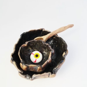 May include: A black ceramic bowl with a rough, textured surface. The bowl has a smaller bowl inside, and a wooden spoon is resting on the rim. A white eyeball with red veins and a yellow pupil is in the center of the bowl.
