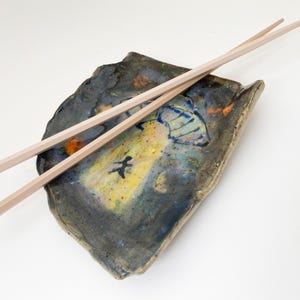 May include: A handmade ceramic chopstick rest with a unique, abstract design. The rest is primarily dark blue and gray, with yellow, orange, and blue accents. Two light-colored wooden chopsticks rest on top.