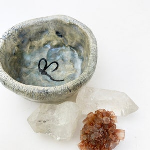 May include: A small, gray ceramic bowl with a black heart design sits on a white surface next to two clear quartz crystals and a cluster of brown selenite crystals.