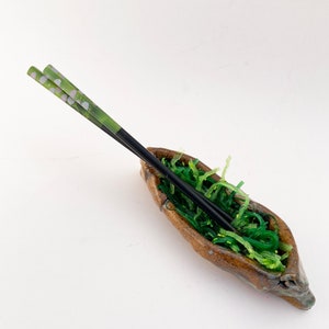 May include: A small, brown ceramic bowl filled with green seaweed and two black chopsticks with green and white floral designs.