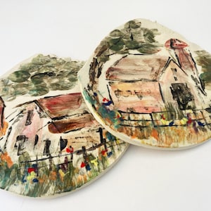 May include: Two ceramic plates with hand-painted scenes of houses and trees. The plates are white with brown, red, pink, green, and blue paint. The plates are oval shaped and have a rustic, folk art style.