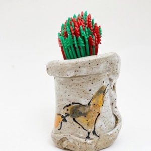 May include: A speckled ceramic container with a hand-painted bird design, holding a bundle of red and green tree-shaped toothpicks. The container has a rustic, handmade appearance.