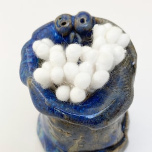 May include: A blue ceramic container with a textured surface and white cotton balls inside. The container has two small holes on the top, resembling eyes.
