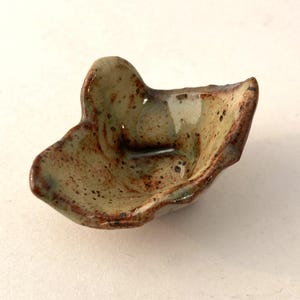 May include: A small, handmade ceramic bowl with an irregular, organic shape. The bowl is glazed in shades of brown, green, and white, with a speckled texture.