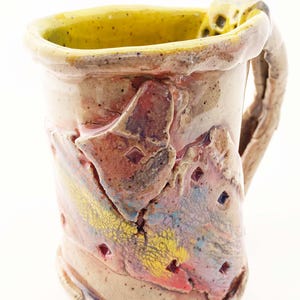 May include: Handmade ceramic mug with a textured surface. The mug features a yellow interior and a handle. The exterior has colorful abstract designs in red, yellow, and blue. The mug is approximately 4 inches tall.